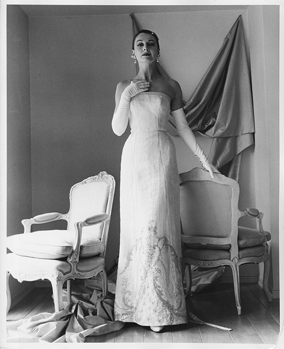 1956 promotional photograph for Galanos. A woman wears a white, beaded, strapless gown with long white opera gloves